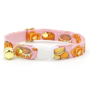 Könnte beinhalten: Rosafarbenes Katzenhalsband mit einem Muster aus Backwaren, darunter Croissants und Zimtschnecken. Das Halsband hat eine weiße Kunststoffschnalle, eine goldfarbene Glocke und eine goldfarbene Metallschlaufe.