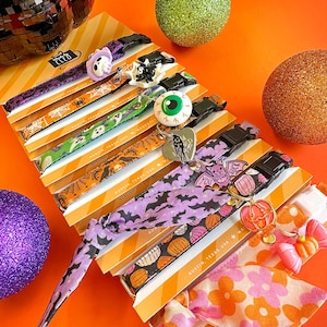 May include: A collection of pet collars featuring Halloween-themed patterns including bats, pumpkins, ghosts, and spiders. Each collar is attached to a branded card and includes a decorative charm such as an eyeball, a bat, or a pumpkin.