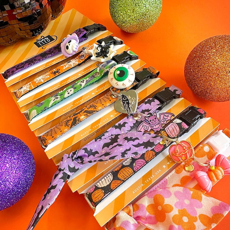 May include: A collection of pet collars featuring Halloween-themed patterns including bats, pumpkins, ghosts, and spiders. Each collar is attached to a branded card and includes a decorative charm such as an eyeball, a bat, or a pumpkin.