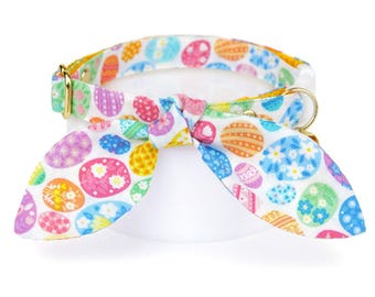 May include: A colorful Easter-themed pet collar with a matching bow tie. The collar and bow tie feature a white background with various pastel-colored Easter egg designs. The collar has a gold-colored metal buckle and a D-ring.