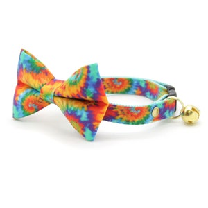 Może przedstawiać: Kolorowy kotek z krawatem w stylu tie-dye z dopasowaną muszką i złotym dzwonkiem.