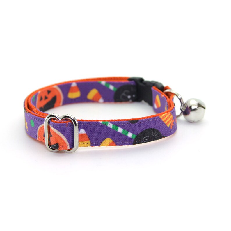 May include: A purple and orange cat collar with a Halloween theme. The collar features a black cat, pumpkins, candy corn, and a bell.