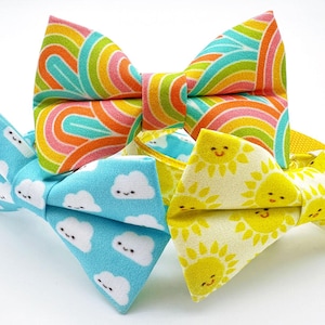 May include: Close-up of three colorful bow ties. One bow tie features a rainbow pattern, another has a light blue background with smiling cloud graphics, and the third has a yellow background with smiling sun graphics. The bow ties are made of fabric.