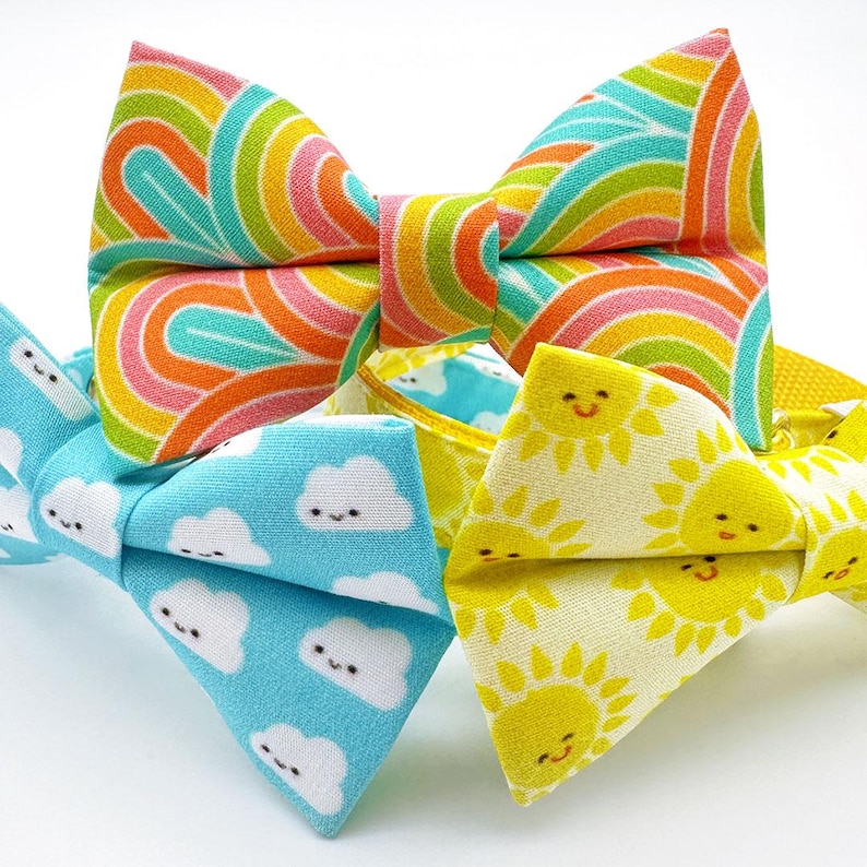 May include: Close-up of three colorful bow ties. One bow tie features a rainbow pattern, another has a light blue background with smiling cloud graphics, and the third has a yellow background with smiling sun graphics. The bow ties are made of fabric.
