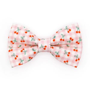 May include: A pink and white gingham bow tie with a repeating pattern of red cherries and green leaves. The bow tie is a classic shape, perfect for adding a touch of whimsy to any outfit.