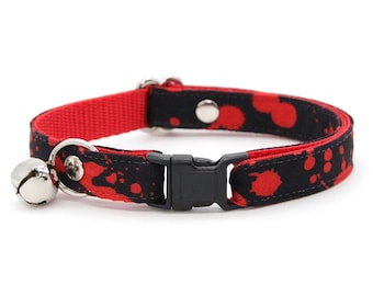 May include: A pet collar featuring a black fabric strap with a red splatter pattern and a solid red inner lining. The collar includes a black plastic side-release buckle, a silver metal D-ring, silver rivets, and a small silver bell attached.