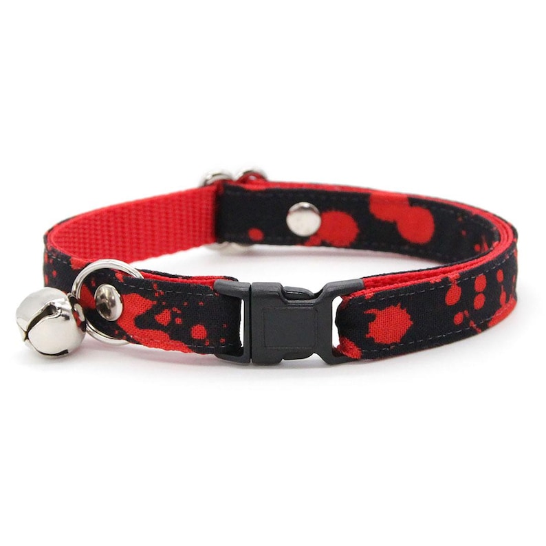 May include: A pet collar featuring a black fabric strap with a red splatter pattern and a solid red inner lining. The collar includes a black plastic side-release buckle, a silver metal D-ring, silver rivets, and a small silver bell attached.