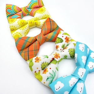 May include: A collection of colorful bow ties with various patterns. Designs include a rainbow stripe, smiling suns, orange plaid, cacti and flowers, and blue clouds with smiling faces. The bow ties are arranged on a white background.