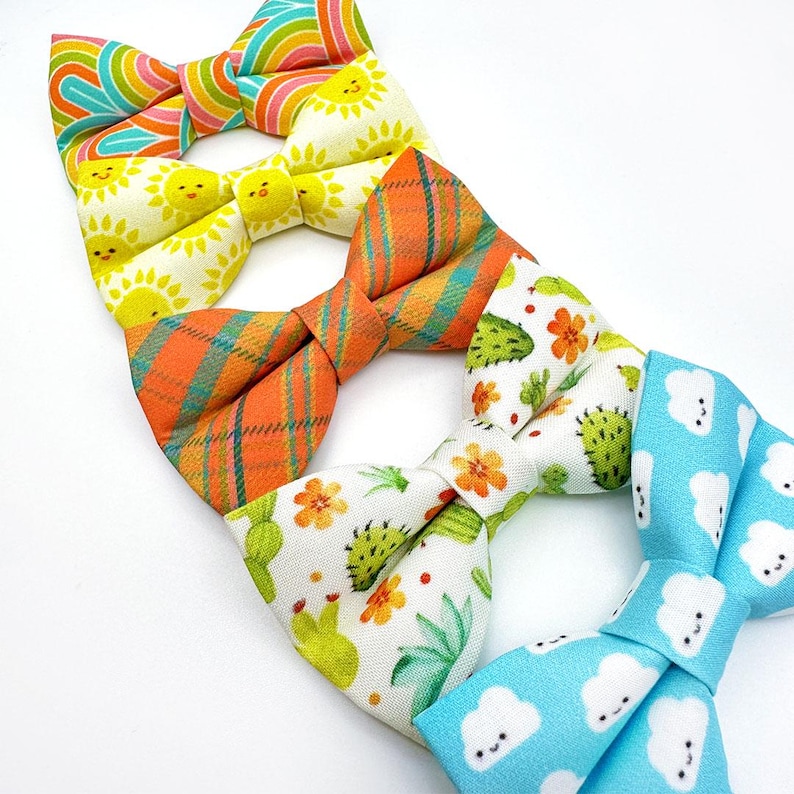 May include: A collection of colorful bow ties with various patterns. Designs include a rainbow stripe, smiling suns, orange plaid, cacti and flowers, and blue clouds with smiling faces. The bow ties are arranged on a white background.