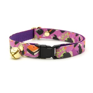 May include: Purple cat collar with a black cat and book print. The collar has a gold bell and a black plastic buckle.
