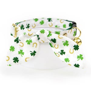 May include: White cat collar with a green shamrock and gold horseshoe pattern, featuring a matching bow tie. The collar has a gold-colored bell and hardware, perfect for St. Patrick's Day.