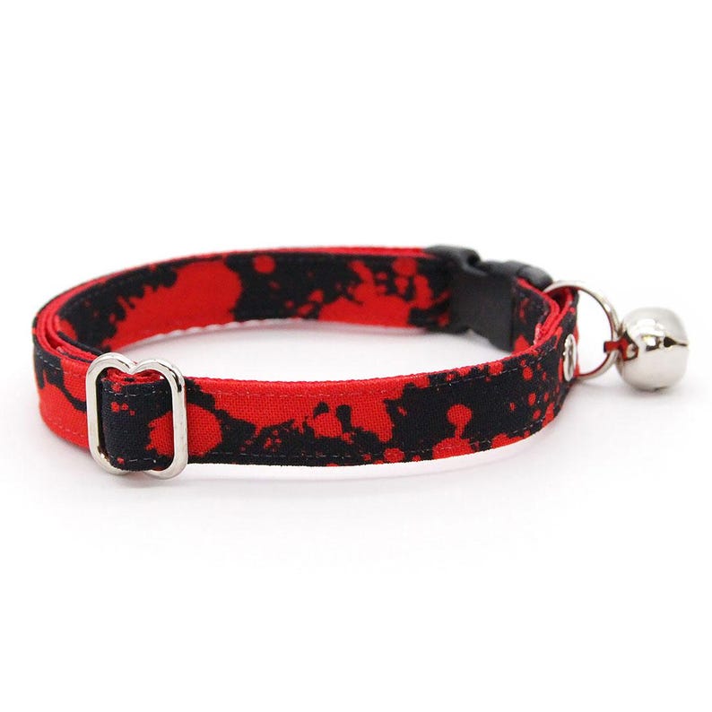 Op de afbeelding: Een halsband voor huisdieren met een rood en zwart inktspatpatroon. De halsband is voorzien van een zwarte plastic klikgesp, een zilverkleurige metalen verstelschuif, een metalen D-ring en een kleine zilveren bel die aan de ring hangt.