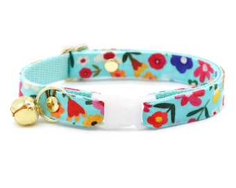 Könnte beinhalten: Ein türkisblaues Katzenhalsband mit Blumenmuster und einer goldenen Glocke. Das Halsband hat eine weiße Kunststoffschnalle.