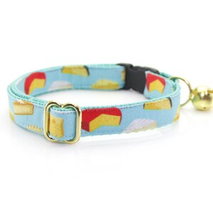 May include: A light blue cat collar with a pattern of colorful cheese wedges. The collar has a gold buckle and a small gold bell.