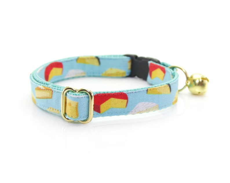 May include: A light blue cat collar with a pattern of colorful cheese wedges. The collar has a gold buckle and a small gold bell.