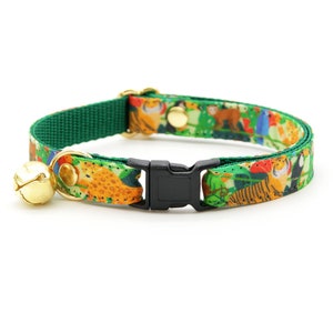 Op de afbeelding: Een groene kattenhalsband met een jungledierenprint en een gouden bel. De halsband heeft een zwarte plastic gesp.