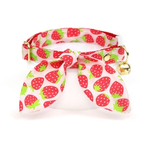 May include: A cat collar with a red and green strawberry pattern on a light pink background. The collar has a bow tie and a gold-colored bell. The collar is made of fabric and has a gold-colored buckle.