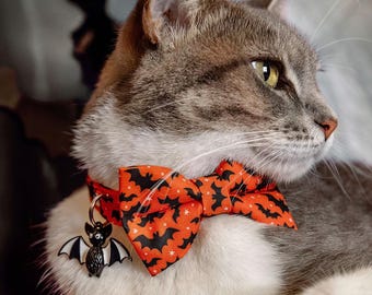 May include: A cat collar featuring an orange fabric bow tie with a black bat and white star pattern. The collar includes a metal ring attached to a small, black and white enamel bat charm that hangs down from the front of the collar band.