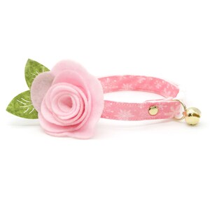May include: Pink cat collar with a pink felt rose and green felt leaves. The collar has a white snowflake pattern and a gold bell.