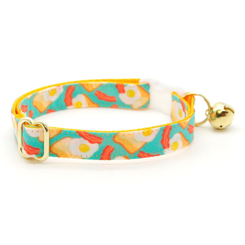 K&ouml;nnte beinhalten: Ein t&uuml;rkisblaues Katzenhalsband mit gelbem Rand und einem Muster aus Spiegeleiern und Speck. Das Halsband hat eine goldene Schnalle und eine goldene Glocke.