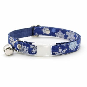 May include: A blue cat collar with a white snowflake pattern and a silver bell. The collar has a white plastic buckle.