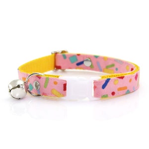 May include: A pink and yellow cat collar with a bell and a white adjustable buckle. The collar has a confetti pattern with colorful geometric shapes.