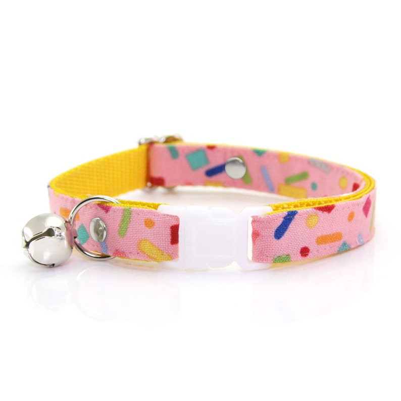 May include: A pink and yellow cat collar with a bell and a white adjustable buckle. The collar has a confetti pattern with colorful geometric shapes.