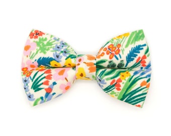 May include: A floral patterned bow tie featuring a cream background with colorful flowers and green leaves. The bow tie has a classic shape and is suitable for various occasions. The fabric has a soft, textured appearance.
