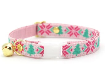 May include: A pink cat collar with a festive pattern of green Christmas trees and red snowflakes. The collar has a white plastic buckle, gold-colored hardware, and a small gold bell.