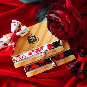 May include: Two pet collars and two matching bow ties featuring red blood splatter patterns on white and black fabric. The collars include black plastic buckles, silver metal D-rings, and small silver bells, all displayed on branded yellow packaging.