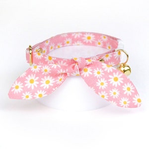 May include: Pink cat collar with a white daisy print and a gold bell. The collar has a bow tie made from the same fabric.