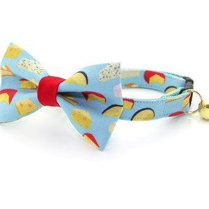 May include: A blue cat collar with a red and white bow tie featuring a cheese print. The collar has a bell attached.