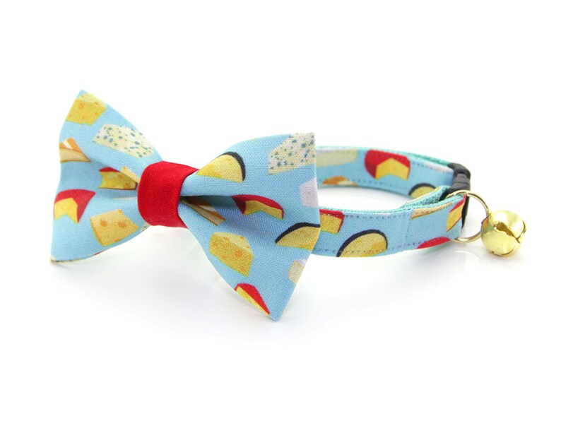 May include: A blue cat collar with a red and white bow tie featuring a cheese print. The collar has a bell attached.