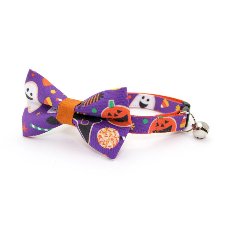 May include: Purple cat collar with a matching bow tie featuring a Halloween print of ghosts, pumpkins, candy corn, and witch hats. The collar has a bell.