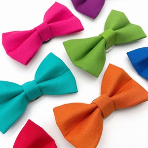 May include: A set of ten colorful bow ties in various colors, including pink, green, turquoise, orange, and red. The bow ties are made of fabric and have a classic bow tie shape.