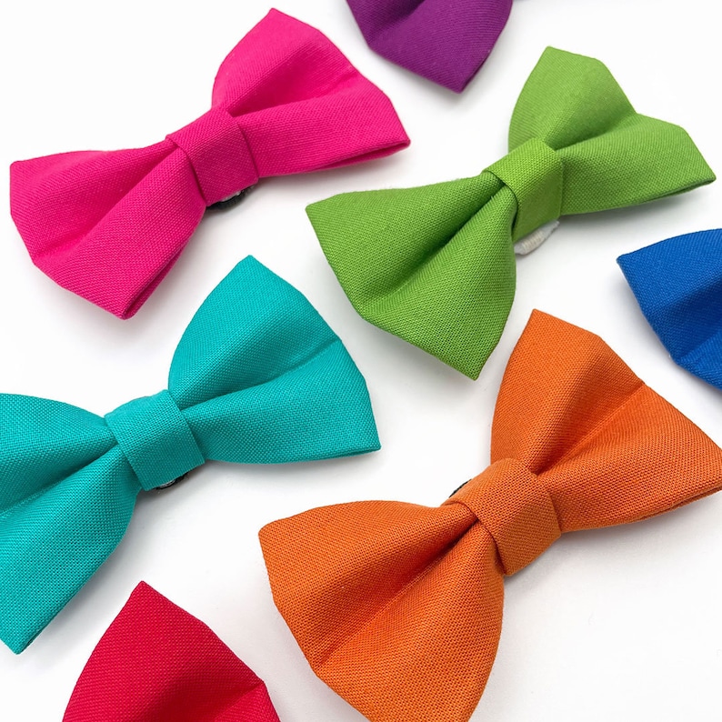 May include: A set of ten colorful bow ties in various colors, including pink, green, turquoise, orange, and red. The bow ties are made of fabric and have a classic bow tie shape.