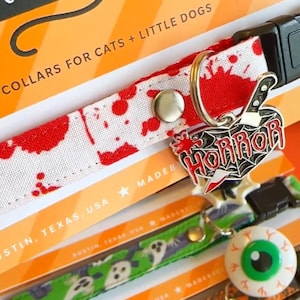 May include: A white pet collar with a red blood splatter pattern featuring a silver-toned metal charm shaped like a heart with a knife and the word HORROR in red text. The collar includes a black plastic buckle and a silver metal D-ring.