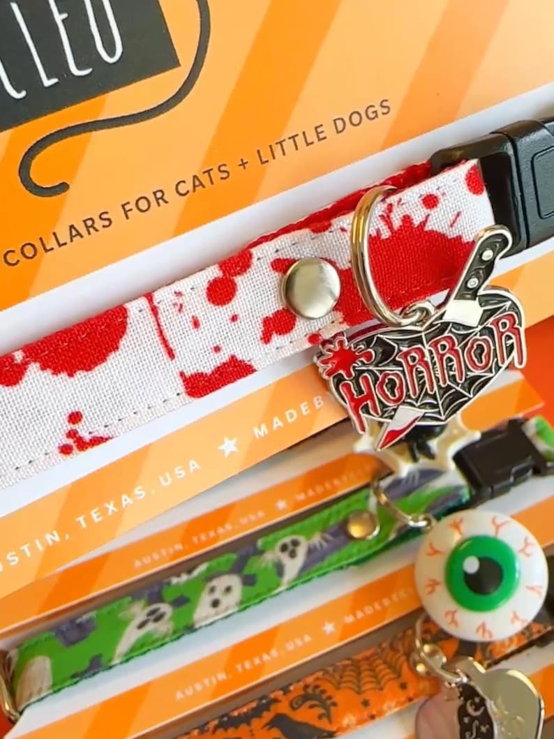 May include: A white pet collar with a red blood splatter pattern featuring a silver-toned metal charm shaped like a heart with a knife and the word HORROR in red text. The collar includes a black plastic buckle and a silver metal D-ring.