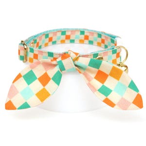 K&ouml;nnte beinhalten: Ein farbenfrohes Haustierhalsband mit passender Schleife. Das Halsband und die Schleife haben ein kariertes Muster in Orange, Gr&uuml;n, Rosa und Blau auf cremefarbenem Hintergrund. Das Halsband hat goldfarbene Hardware.