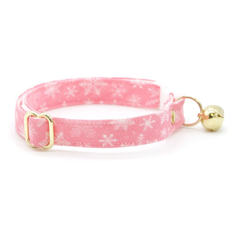 May include: Pink cat collar with a white snowflake pattern and a gold bell.
