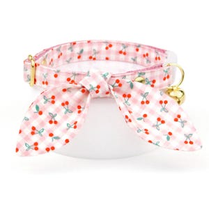 May include: A pink and white gingham cat collar with a cherry pattern and a matching bow. The collar has a gold-colored metal buckle and a small gold bell. The bow is tied in the center of the collar.