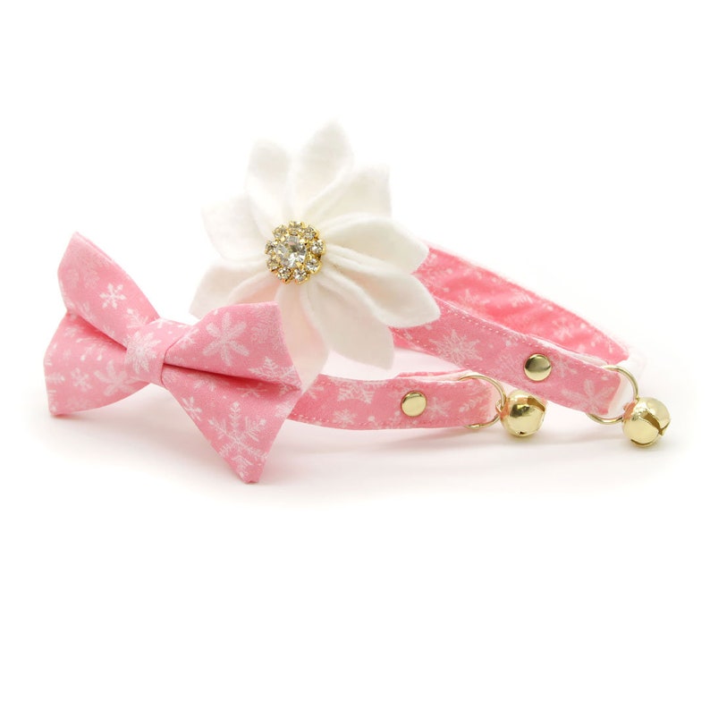 May include: Pink snowflake patterned cat collar with a matching bow tie and a white flower with a rhinestone center.