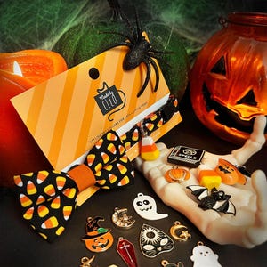 May include: A black pet collar featuring a candy corn pattern and a matching bow tie, attached to a branded orange and white striped display card. The set includes a candy corn charm and various Halloween-themed metal charms like ghosts, bats, and pumpkins.