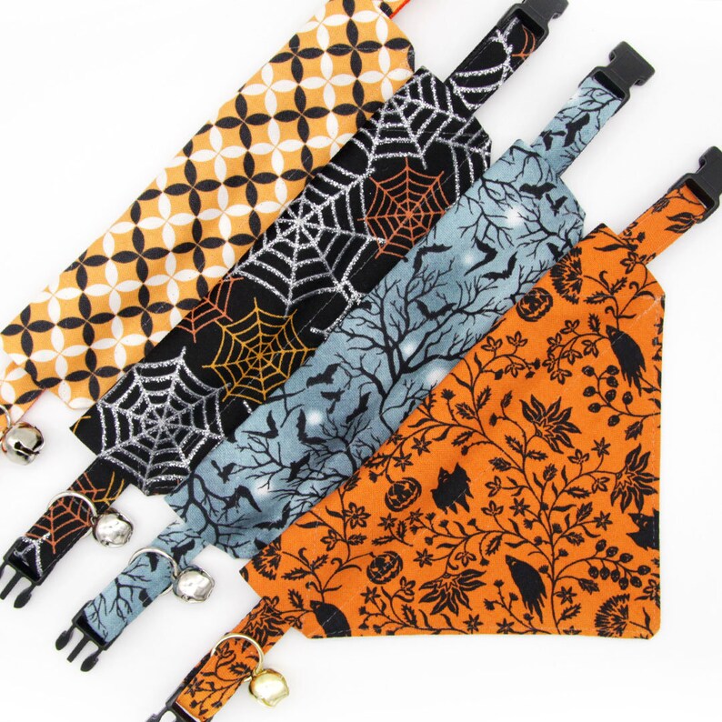 May include: Four different Halloween-themed cat collars with bells. The collars are made of fabric with various patterns, including orange with black floral designs, black with silver glitter and spiderwebs, blue with white bats and stars, and orange with black geometric shapes.