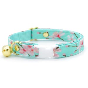 May include: A turquoise cat collar with a floral pattern of pink blossoms. The collar has a white plastic buckle, a gold-colored bell, and gold-colored metal hardware. The collar is designed for a cat.