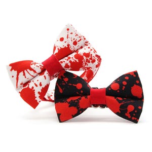 May include: Two bow ties with a red splatter pattern. One bow tie features a white base with red paint-like splatters, while the other bow tie has a black base with red paint-like splatters. Both bow ties have a solid red center knot.