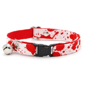 Op de afbeelding: Een witte halsband voor huisdieren met een rood spetterpatroon, voorzien van een zwarte plastic gesp, een zilveren metalen ring en een kleine zilveren bel. De halsband is gemaakt van stof met een rode voering aan de binnenzijde.
