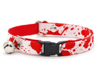 May include: A white pet collar featuring a red splatter pattern, constructed with a black plastic side-release buckle, a silver metal D-ring, and a small silver bell attached to the side. The collar is made of fabric with a red interior lining.