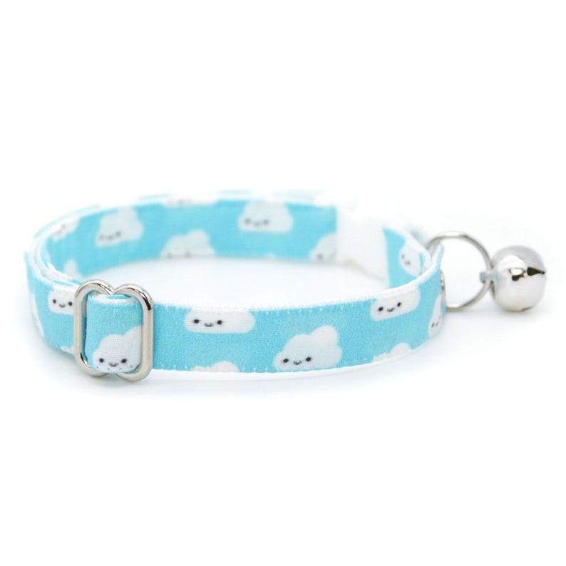 May include: A light blue cat collar with a repeating pattern of smiling white clouds. The collar has a silver-colored metal buckle and a small silver bell attached. The collar is designed for a cat.