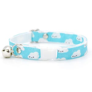 May include: A light blue cat collar with a white cloud pattern and a silver bell. The collar has a white plastic buckle and a metal loop for attaching a leash. The clouds have smiling faces.